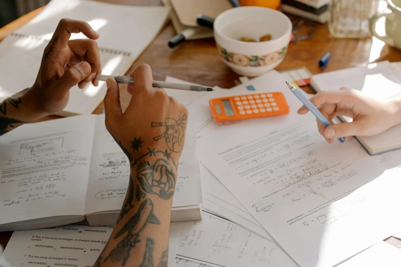 a person sitting at a table with papers and a calculator, by Sam Charles, pexels, process art, arms covered in gang tattoo, coworkers, taken on iphone 1 3 pro, wooden desks with books