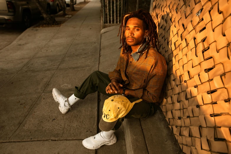 a man with dreadlocks sitting against a brick wall, by Kurt Trampedach, pexels contest winner, photorealism, yellow hue, kara walker james jean, casual pose, sidewalk