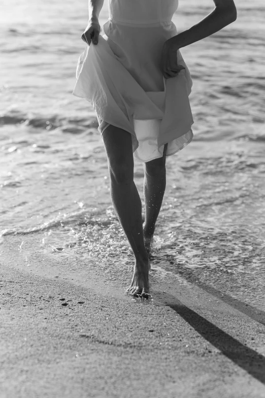 a black and white photo of a woman walking on the beach, inspired by Max Dupain, unsplash, renaissance, beautiful legs, wearing a white sundress, nymph in the water, ( ( photograph ) )