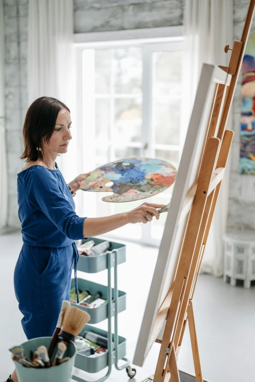 a woman in a blue dress painting on an easel, a painting, inspired by artist, pexels contest winner, in a white boho style studio, trending on artlist, artist wearing overalls, profile image