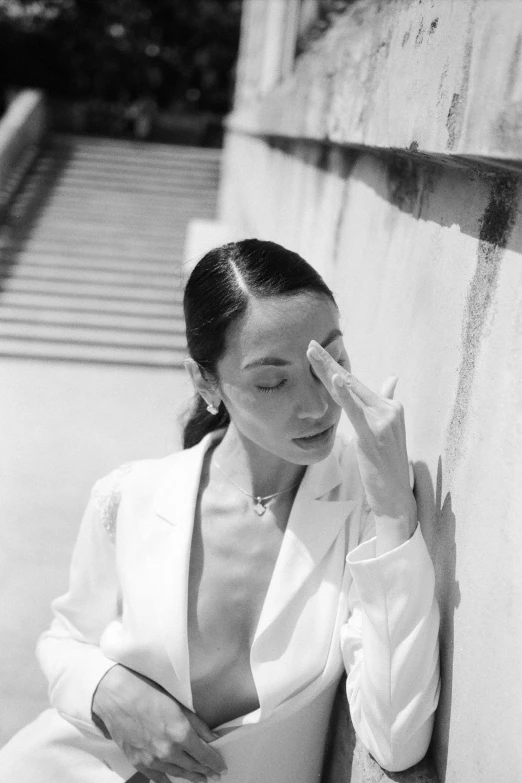 a black and white photo of a woman leaning against a wall, inspired by Marina Abramović, kiko mizuhara, wearing a white tuxedo, blinding sun, wearing off - white style