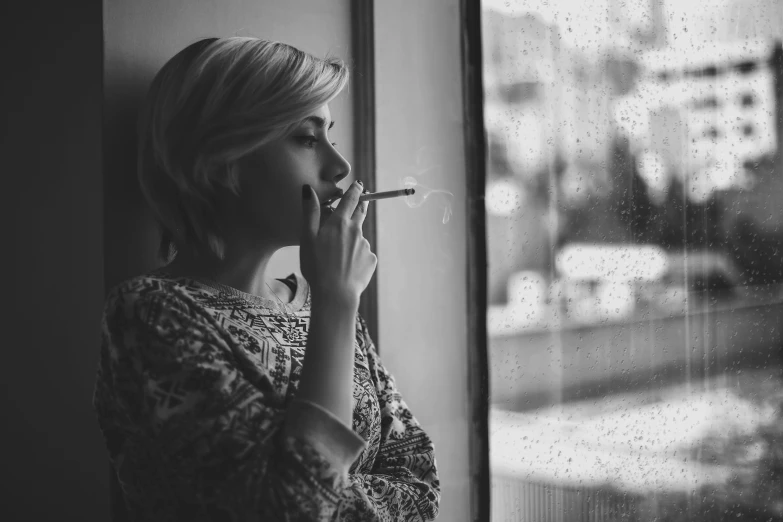 a woman smoking a cigarette in front of a window, a black and white photo, inspired by Elsa Bleda, pexels, young blonde woman, realistic »