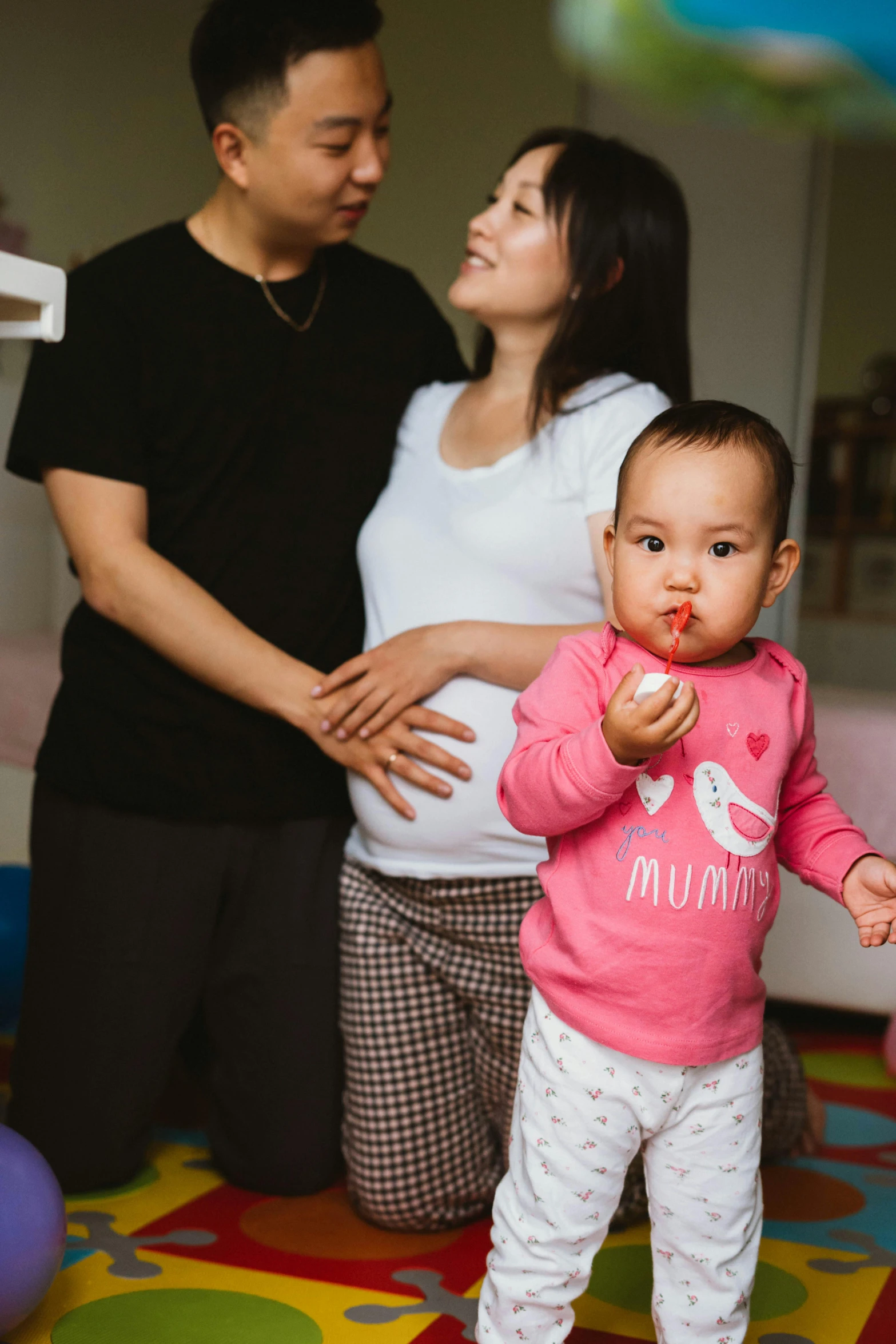 a man standing next to a woman holding a baby, pexels contest winner, young asian girl, at home, pink, square