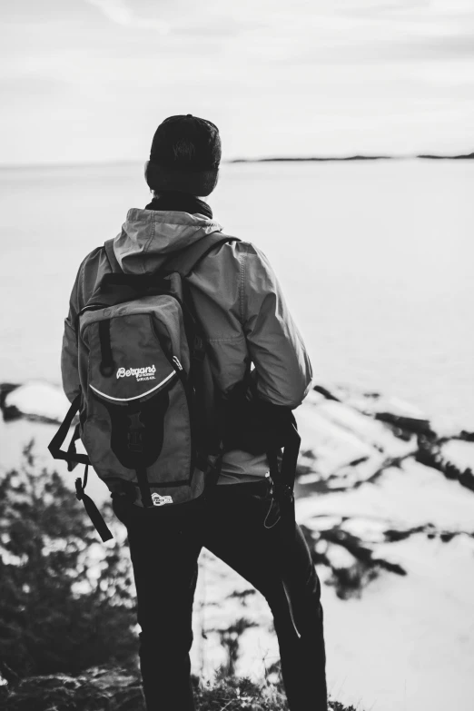 a man standing on the edge of a cliff overlooking a body of water, a black and white photo, inspired by Daniel Ljunggren, unsplash, backpack, ✨🕌🌙, techwear clothes, profile picture 1024px