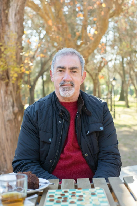 a man sitting at a table with food and drinks, ana de la reguera portrait, trees in the background, portrait image, 5 5 yo