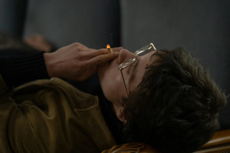 a man laying on a couch with a cigarette in his mouth, by Attila Meszlenyi, trending on pexels, finn wolfhard, fire lit, jewish young man with glasses, praying with tobacco