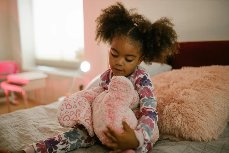 a little girl sitting on a bed holding a teddy bear, by Alice Mason, pexels, african american girl, in the bedroom at a sleepover, multicoloured, hugging