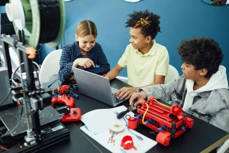 a group of children sitting around a table with a laptop, red and black robotic parts, steam workshop, gettyimages, keter class