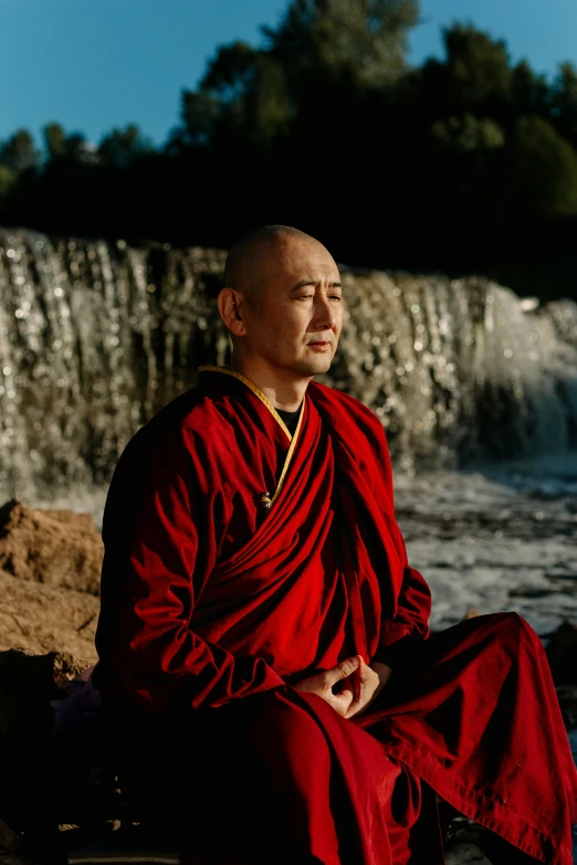 a man in a red robe sitting in front of a waterfall, a portrait, inspired by Zhao Mengfu, unsplash, frans lanting, evening light, new mexico, monk clothes