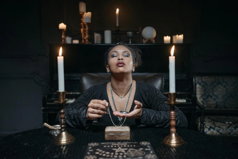 a woman sitting at a table surrounded by candles, a portrait, trending on pexels, magical realism, occult jewelry, afrocentric mysticism, holding nevermore, on the altar