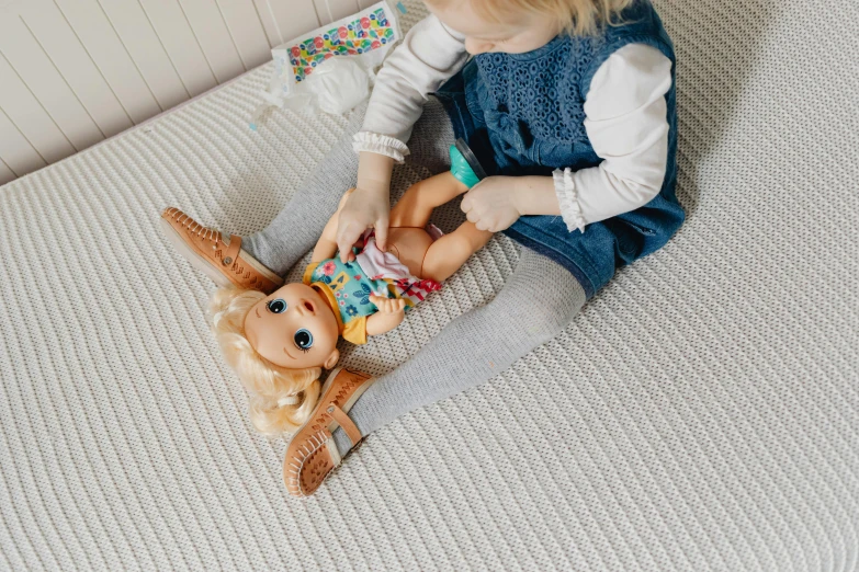 a little girl sitting on top of a bed next to a doll, inspired by Myles Birket Foster, unsplash, top angle view, elsa frozen, multicoloured, blond