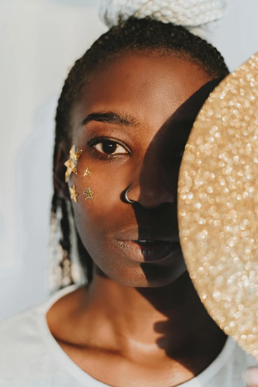 a woman holding a straw hat in front of her face, trending on pexels, afrofuturism, gold eyeshadow, starry, white and gold color palette, looking into a mirror