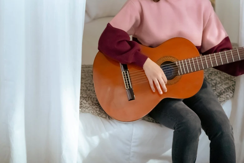 a woman sitting on a bed playing a guitar, inspired by Gwen John, trending on pexels, sweater, teenager girl, brown and pink color scheme, sitting on an armchair