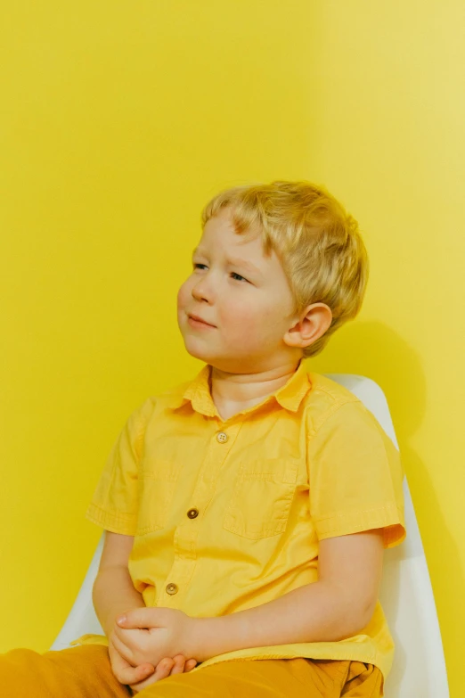 a young boy sitting on top of a white chair, yellow backdrop, wearing a shirt, looking off to the side, hansa yellow