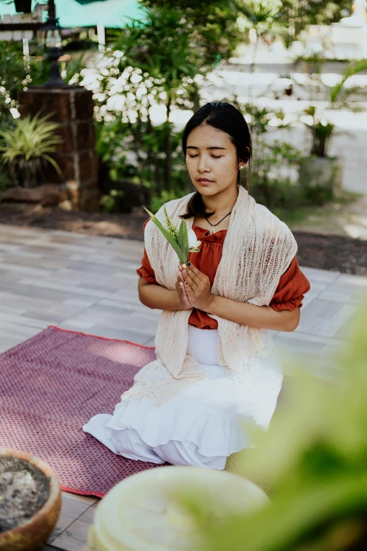 a woman sitting on a mat in front of a potted plant, inspired by Ruth Jên, unsplash, renaissance, sukhothai costume, praying, white and pink cloth, casting a protection spell