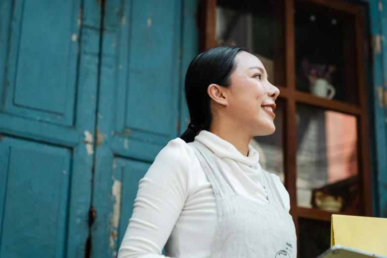 a woman standing in front of a blue door, a portrait, inspired by Cui Bai, pexels contest winner, happening, wearing an apron, looking upwards, profile image, stood outside a corner shop