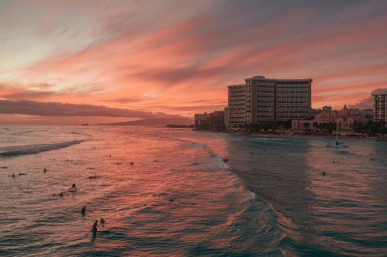 surfers in the ocean at sunset with buildings in the background, by Daniel Lieske, pexels contest winner, pink and grey clouds, slightly tanned, hawaii, red hues