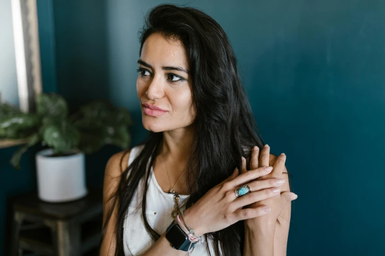 a woman standing in front of a blue wall, a portrait, by Kristian Zahrtmann, pexels contest winner, hurufiyya, wearing two metallic rings, messy long black hair, sitting on a mocha-colored table, turquoise jewelry