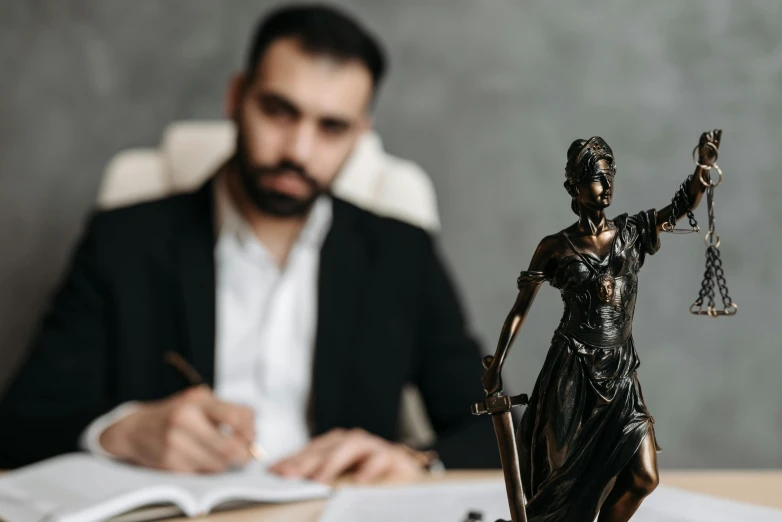 a man sitting at a desk with a statue of lady justice, a statue, by Adam Marczyński, pexels contest winner, kurdish lawyer, man holding spear, thumbnail, high quality image