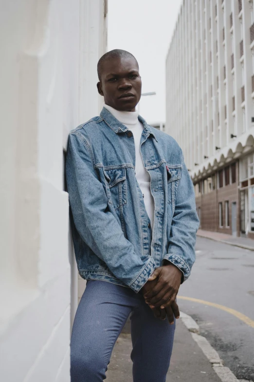 a man in a denim jacket leaning against a wall, an album cover, inspired by Theo Constanté, pexels contest winner, style of seb mckinnon, standing in a city center, tight wrinkled cloath, riyahd cassiem
