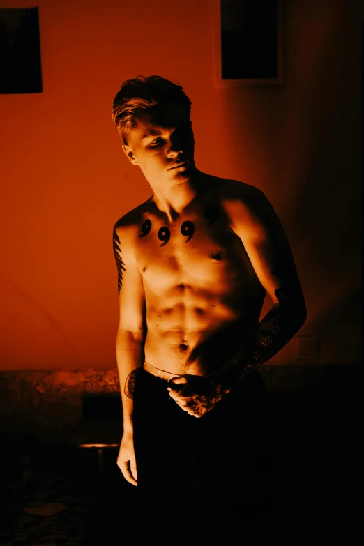 a shirtless man standing in front of a red wall, a tattoo, inspired by Nikolaj Abraham Abildgaard, lit up in a dark room, alternate album cover, 18 years old, lean man with light tan skin