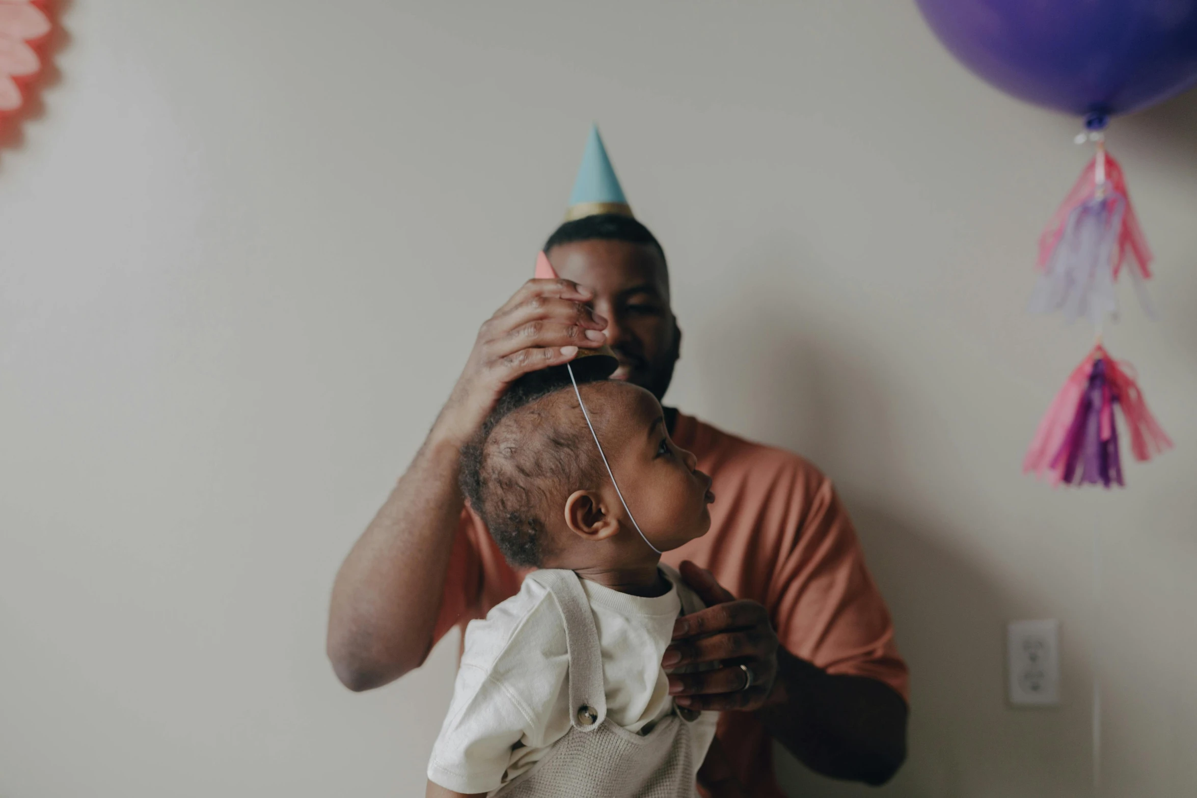 a man holding a baby wearing a party hat, pexels contest winner, dark skinned, happy birthday, growth on head, party balloons