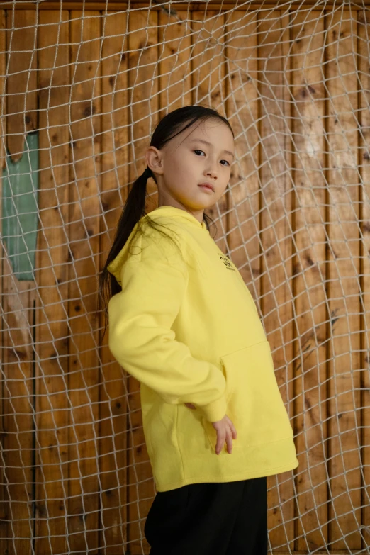 a little girl standing in front of a net, inspired by Tang Di, pexels contest winner, happening, wearing a yellow hoodie, outlive streetwear collection, posed in profile, modeling for dulce and gabanna