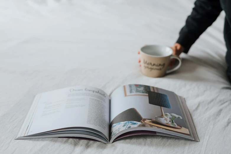 an open book sitting on top of a bed next to a cup of coffee, by Harvey Quaytman, happening, homes and garden magazine, lined in cotton, casually dressed, medium level shot