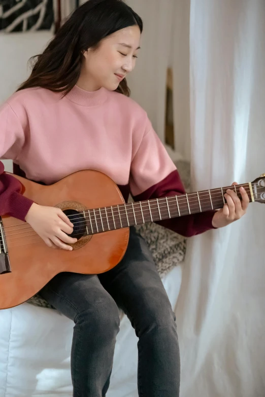 a woman sitting on a bed playing a guitar, inspired by Ni Yuanlu, trending on pexels, wearing casual sweater, brown and pink color scheme, for kids, wearing sweatshirt