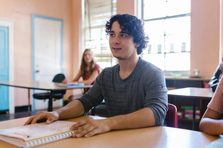 a man sitting at a table with a notebook in front of him, pexels contest winner, academic art, standing in class, lachlan bailey, emma uber, looking across the shoulder