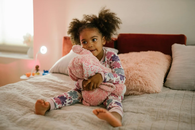 a little girl sitting on a bed holding a teddy bear, inspired by Myles Birket Foster, pexels, african american, wearing pajamas, pink iconic character, paisley