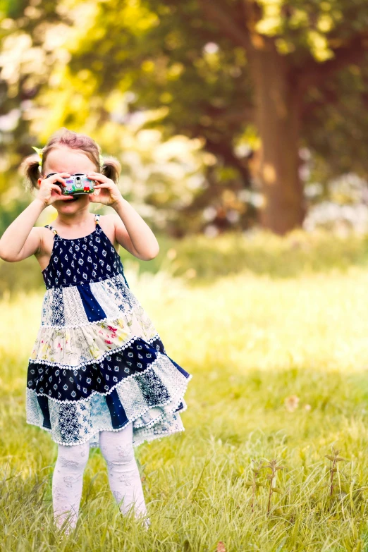 a little girl standing on top of a lush green field, a picture, shutterstock, binoculars, blue checkerboard dress, at a park, patterned clothing