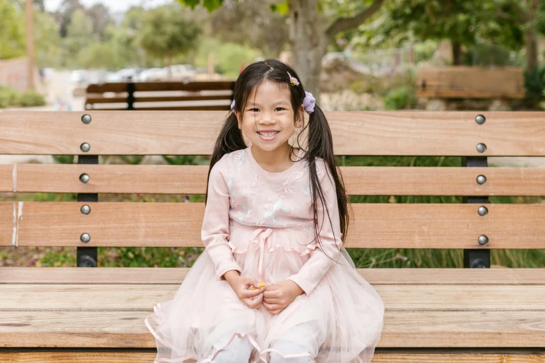 a little girl sitting on top of a wooden bench, wearing a pink dress, jia ruan, manuka, she is smiling