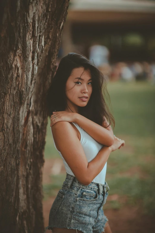 a woman leaning against a tree in a park, by Adam Marczyński, pexels contest winner, realism, tan skin a tee shirt and shorts, beautiful young asian woman, sydney park, summer evening