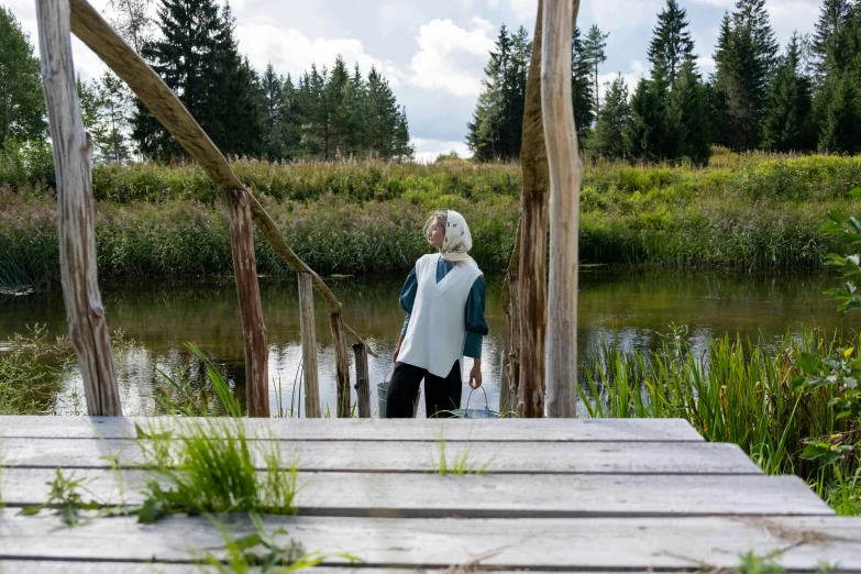 a woman standing on a dock next to a body of water, by Grytė Pintukaitė, hurufiyya, in the countryside, wearing a vest top, artisanal art, blue tunic and robes