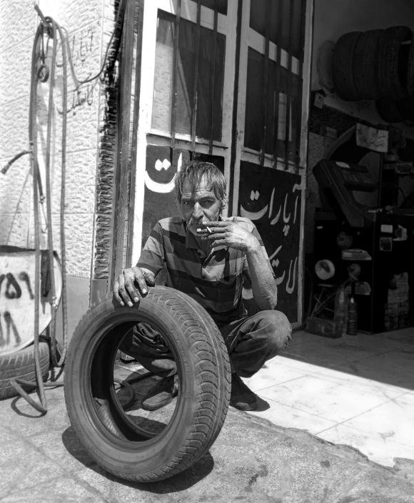 a man sitting on the ground next to a tire, by Youssef Howayek, garage, jerusalem, smoking, with an intricate