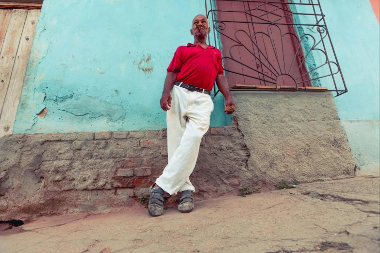 a man standing in front of a blue building, by William Berra, pexels contest winner, an old lady with red skin, built on a steep hill, gogo : :, blue long pants and red shoes