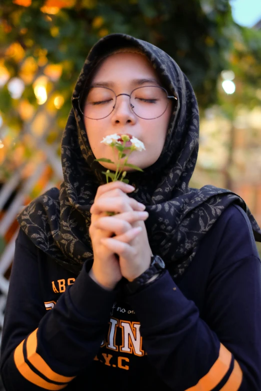 a woman holding a flower in front of her face, a picture, by Maryam Hashemi, pexels contest winner, hurufiyya, wearing small round glasses, ☁🌪🌙👩🏾, hooded, at college