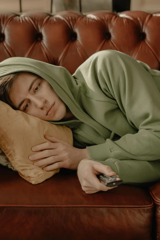 a person laying on a couch with a remote, a colorized photo, by Mark Arian, trending on pexels, hyperrealism, beige hoodie, concerned expression, green clothes, joe keery