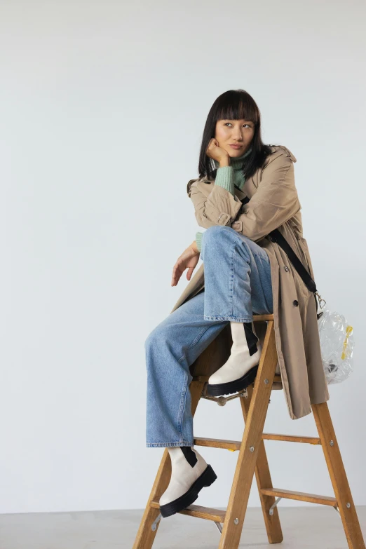 a woman sitting on top of a wooden ladder, wearing a long beige trench coat, roots and hay coat, gemma chan, banner