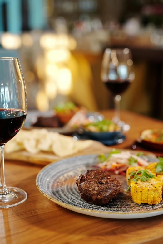 a wooden table topped with plates of food and a glass of wine, holding a glass of red wine, patagonian, luxury, beef