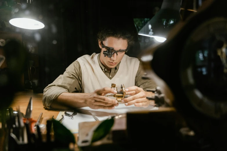 a man sitting at a table working on something, by Adam Marczyński, pexels contest winner, jewelry engraved in scarab, wearing victorian brass goggles, low-light, student