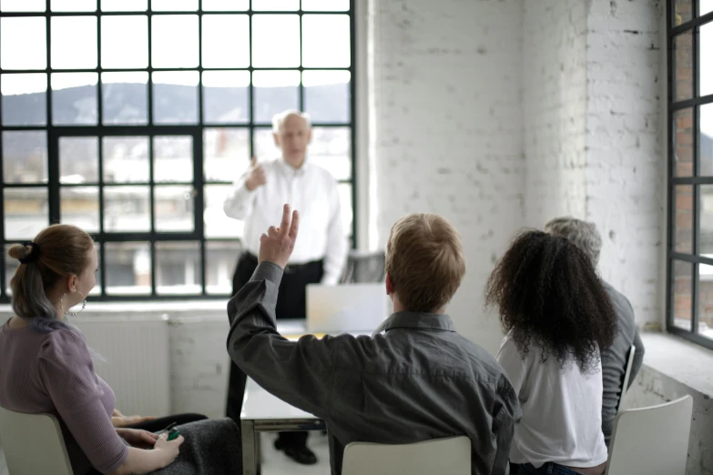 a group of people sitting around a table in front of a window, indistinct man with his hand up, standing in class, listing image, thumbnail