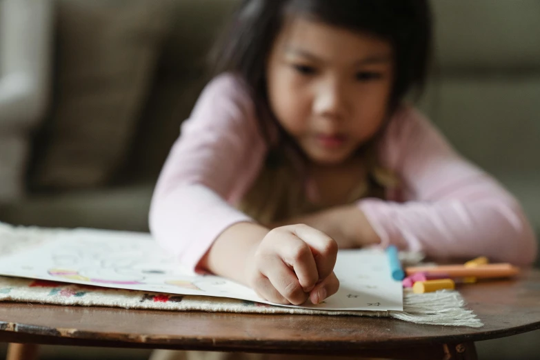 a little girl sitting at a table writing on a piece of paper, a child's drawing, by Lee Loughridge, pexels contest winner, young asian girl, closeup - view, reading a book, full colour