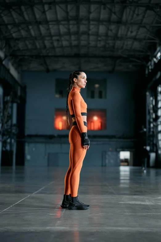 a woman in an orange jumpsuit standing in a warehouse, inspired by Caro Niederer, antipodeans, cyborg tech on body and legs, wearing track and field suit, full body profile pose, promo image