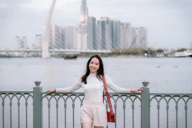 a woman standing on a bridge with a city in the background, a picture, inspired by Ruth Jên, pexels contest winner, in front of white back drop, pretty face sharp chine, at the waterside, mai anh tran