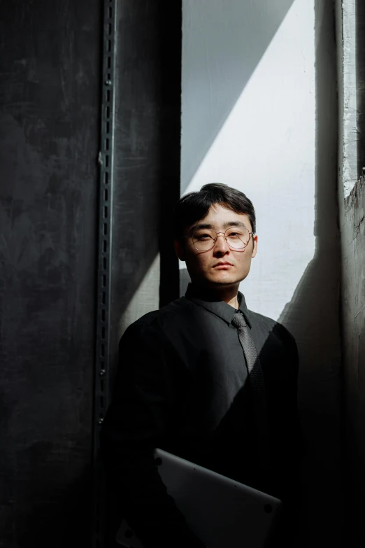 a man standing next to a wall with a laptop, an album cover, inspired by Joong Keun Lee, wearing a black shirt, a portrait of issey miyake, conan, in a black room