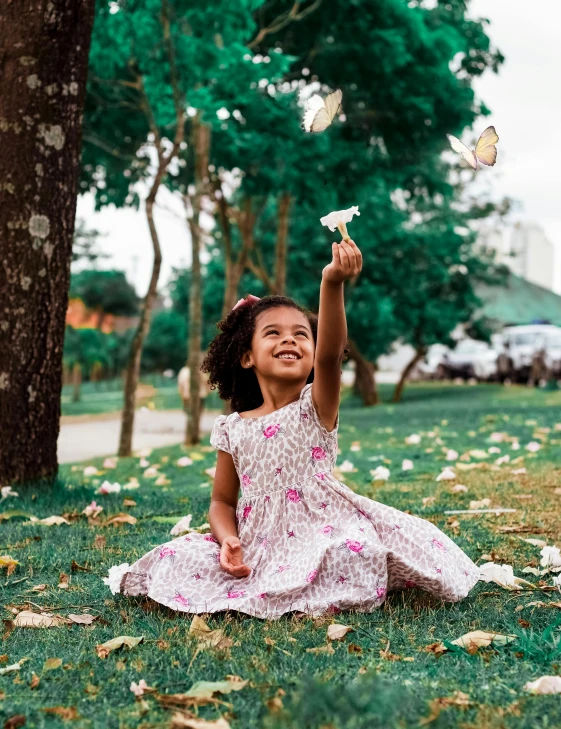 a little girl that is sitting in the grass, pexels contest winner, butterflies flying, with a tree in the background, 🌸 🌼 💮, mai anh tran