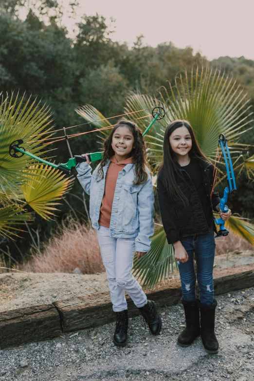 a couple of girls standing next to each other, by Jessie Algie, pexels contest winner, hurufiyya, fronds, wielding a bow, mini model, family friendly