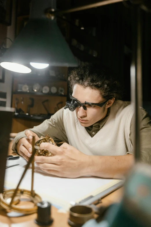 a man sitting at a table working on something, by Everett Warner, trending on pexels, renaissance, wearing victorian brass goggles, jewelry engraved in scarab, lit up, student